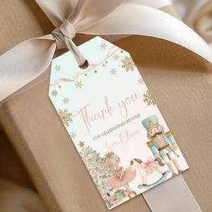 May include: A rectangular gift tag with a light blue background, gold snowflakes, and pink ribbon accents. It features a ballerina, a nutcracker, and a rocking horse, with the text Thank you FOR CELEBRATING WITH ME love, Olivia.