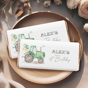 May include: Two white candy bar wrappers with a green tractor illustration and the text "ALEX'S 1st Birthday" and "ALEX'S Birthday". The wrappers are on a wooden tray with a floral background.