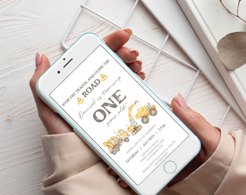 May include: A smartphone displays a birthday invitation with a construction theme. The invitation features illustrations of construction vehicles, the text "STOP THE TRAFFIC AND CLOSE THE ROAD," and the words "ONE year old!" in a playful font. The invitation includes event details.