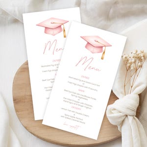 Op de afbeelding: Twee menukaarten met een roze afstudeerhoed illustratie en gouden kwast. Het menu bevat voorgerechten, hoofdgerechten en desserts. De kaarten liggen op een houten dienblad met een witte servet en droogbloemen.
