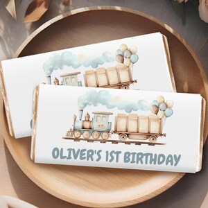 Op de afbeelding: Twee witte chocoladeverpakkingen met een blauwe trein en bruine goederenwagons, blauwe en witte ballonnen en de tekst "Oliver's 1st Birthday".