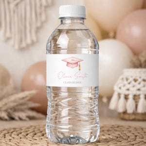 May include: Clear plastic water bottle with a white label featuring a pink graduation cap and the text "Olivia Smith CLASS OF 20XX". The bottle has a white cap and is filled with clear liquid.