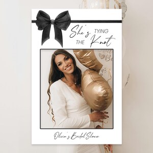 May include: A white photo booth frame with a black bow and the text "She's Tying the Knot" in black script. The frame has a photo of a woman with blonde hair holding gold balloons. The text "Olivia's Bridal Shower" is at the bottom of the frame.