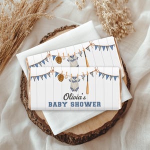 May include: Two rectangular candy bar wrappers featuring a white wood-plank background with a clothesline displaying a baseball glove, ball, jersey with the word Rookie, whistle, and bat. Blue pennant banners hang above. Text reads Olivia's BABY SHOWER.