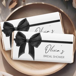 Könnte beinhalten: Zwei weiße Schokoladenriegelverpackungen mit schwarzen Schleifen und dem aufgedruckten Text "Olivia's Bridal Shower".