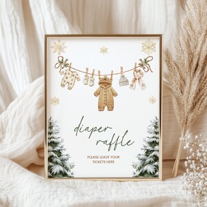 May include: A framed sign with a gold border features a winter-themed baby shower design. The sign includes illustrations of baby clothes hanging on a clothesline, snowflakes, and evergreen trees. The text reads "diaper raffle" and "please leave your tickets here."