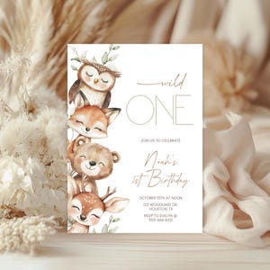 Può includere: Un biglietto d'invito bianco con la scritta "wild ONE" e "Noah's 1st Birthday". Il biglietto presenta illustrazioni di un gufo, una volpe, un orso e un cervo. L'invito include data, ora, luogo e informazioni RSVP.