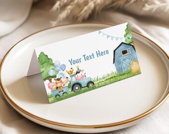 May include: A folded place card with a farm theme. The card features a blue tractor carrying farm animals, a blue barn, and the text "Your Text Here". The card is on a white plate with a gold rim.