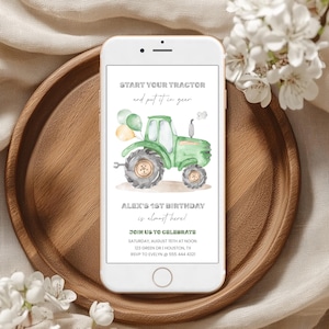 May include: A white smartphone displays a birthday invitation with a green tractor illustration and text. The invitation reads "START YOUR TRACTOR and put it in gear" and "ALEX'S 1ST BIRTHDAY is almost here!" The phone rests on a wooden tray.