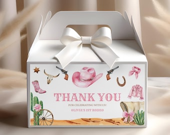 May include: White favor box with a pink and white cowboy themed design. The box has a white ribbon and bow. The box has the text "Thank You" and "Olivia's 1st Rodeo".