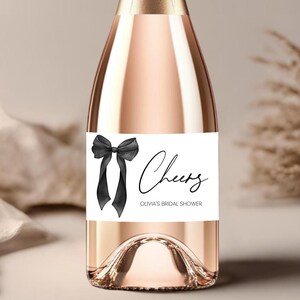 May include: A white wine bottle label with a black bow and the text "Cheers Olivia's Bridal Shower".