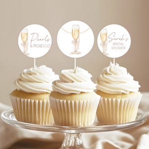 Może przedstawiać: Trzy babeczki z białym lukrem i dekoracyjnymi topperami. Toppery przedstawiają kieliszki do szampana i tekst „Pearls & Prosecco” oraz „Sarah's Bridal Shower”. Babeczki są prezentowane na szklanym stojaku na ciasto.
