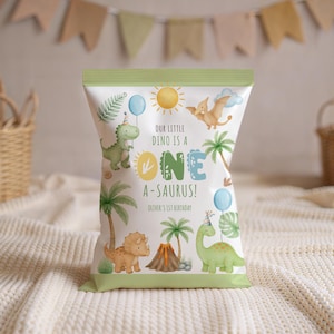 Op de afbeelding: Een witte traktatiezak met een groene bovenkant, met een dinosaurus thema. De zak heeft illustraties van dinosaurussen, palmbomen, een vulkaan en de tekst "OUR LITTLE DINO IS A ONE A-SAURUS!" en "OLIVER'S 1ST BIRTHDAY".