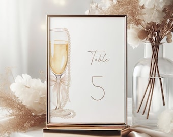 May include: A framed table number card with a champagne glass illustration. The card features the word "Table" and the number "5" in a delicate script. The frame and stand are gold-colored, and the background includes floral arrangements.