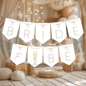 Puede incluir: Una guirnalda blanca con las palabras "BRIDE to BE" en una fuente delicada, ensartada con una guirnalda de perlas y lazos. Una ilustración de acuarela de una copa de champán aparece en una de las pancartas. Decoración para una despedida de soltera.