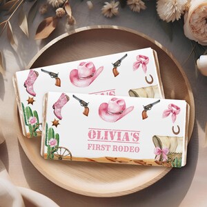 May include: Pink and white personalized chocolate bar wrappers with a western theme. The wrappers feature a pink cowboy hat, boots, a lasso, and a wagon. The text on the wrapper reads "Olivia's First Rodeo".