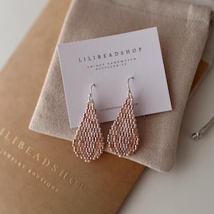 Puede incluir: Un par de aretes delicados en forma de lágrima hechos con pequeñas cuentas de color rosa claro y plata. Los aretes están empaquetados en un sobre pequeño de color marrón con el texto "LILI BEADS SHOP Jewelry Boutique" y una tarjeta blanca con el texto "LILI BEADS SHOP Unique Handwoven Accessories".
