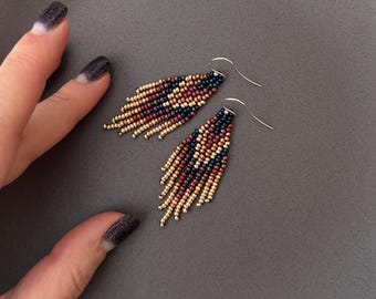 Pode incluir: Um par de brincos de contas com um design de franjas. Os brincos apresentam um padrão de contas azul marinho, bordô e dourado. São exibidos em um prato cinza.