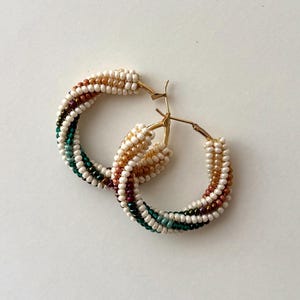 May include: A pair of gold hoop earrings with a beaded design. The beads are arranged in a spiral pattern, featuring colors like white, brown, burgundy, green, and gold. The earrings are set against a white background.