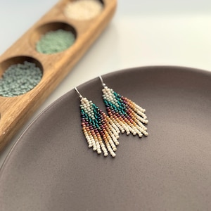 Peut inclure: Une paire de boucles d'oreilles pendantes avec un dégradé de perles turquoise, brunes et blanches. Les boucles d'oreilles sont sur une assiette grise.