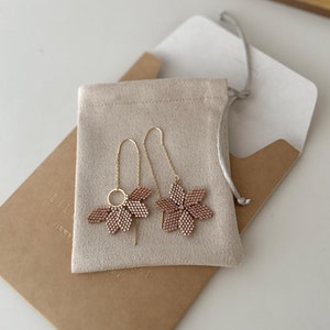 May include: A pair of delicate gold earrings with a flower design made of small brown beads. The earrings are displayed in a small beige pouch.
