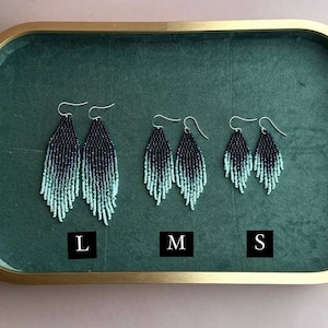 Könnte beinhalten: Perlenohrringe in drei Größen (L, M, S) auf einem grünen Samttablett. Die Ohrringe gehen von Schwarz zu einem hellen Türkis über. Das Tablett hat einen goldfarbenen Rahmen. Der Hintergrund ist hellrosa.