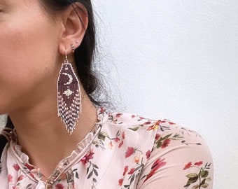 Peut inclure: Une paire de longues boucles d'oreilles en perles avec un motif bordeaux et blanc. Les boucles d'oreilles pendent des oreilles d'une personne portant un chemisier à fleurs.