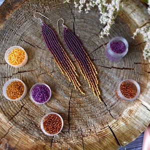 May include: A pair of long, beaded earrings with a gradient of purple and gold beads. The earrings are hanging from silver hooks. The beads are arranged in a cascading pattern, creating a unique and eye-catching design.