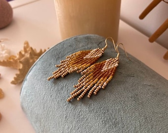 May include: A pair of gold beaded earrings with a fringe design. The earrings are made with small, round beads in shades of gold, brown, and white.