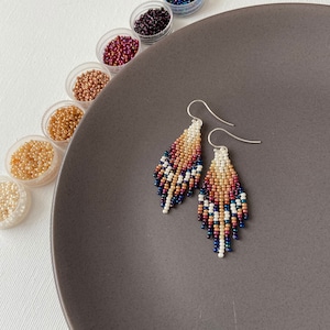 May include: A pair of beaded earrings in a teardrop shape. The earrings are made with small, colourful beads in shades of brown, white, and blue. The earrings are hanging from silver hooks.