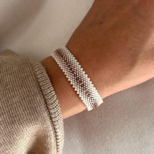 以下が含まれることがあります： 白、茶色、ベージュの色合いのビーズブレスレットが手首に着用されています。 ブレスレットは、小さく均等に配置されたビーズでできており、テクスチャード加工された表面を作り出しています。 ブレスレットのデザインは、交互の色パターンを特徴としています。