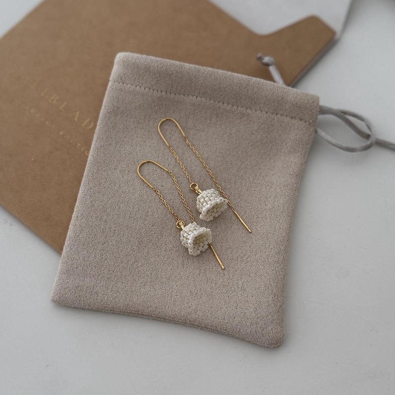 May include: A pair of gold dangle earrings with white beaded bell charms. The earrings are displayed on a beige velvet pouch.