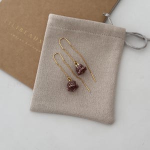 May include: A pair of delicate gold earrings with small, beaded, mauve-colored bell charms hanging from thin chains. The earrings are displayed on a light brown suede pouch.