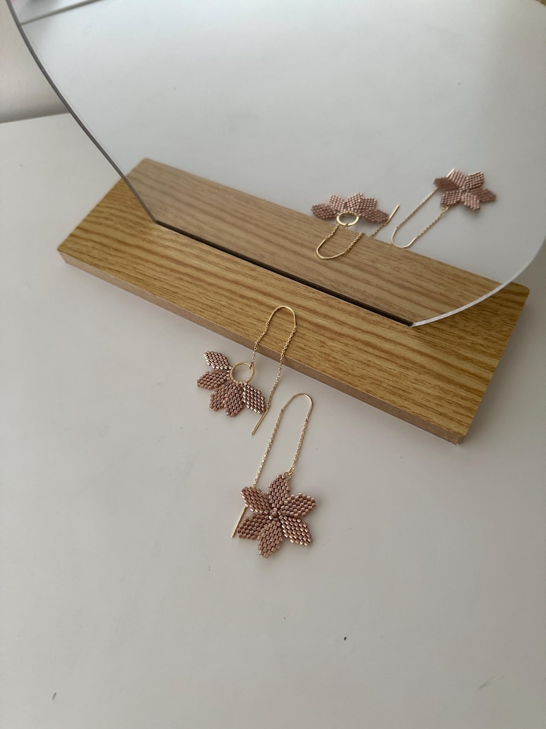 May include: A pair of delicate gold earrings with pink flower-shaped charms. The earrings are hanging from a wooden surface.