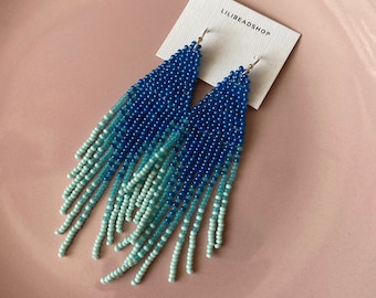 May include: A pair of blue and turquoise beaded fringe earrings with silver hooks. The earrings are displayed on a white card with the text "LILIBEADSHOP".