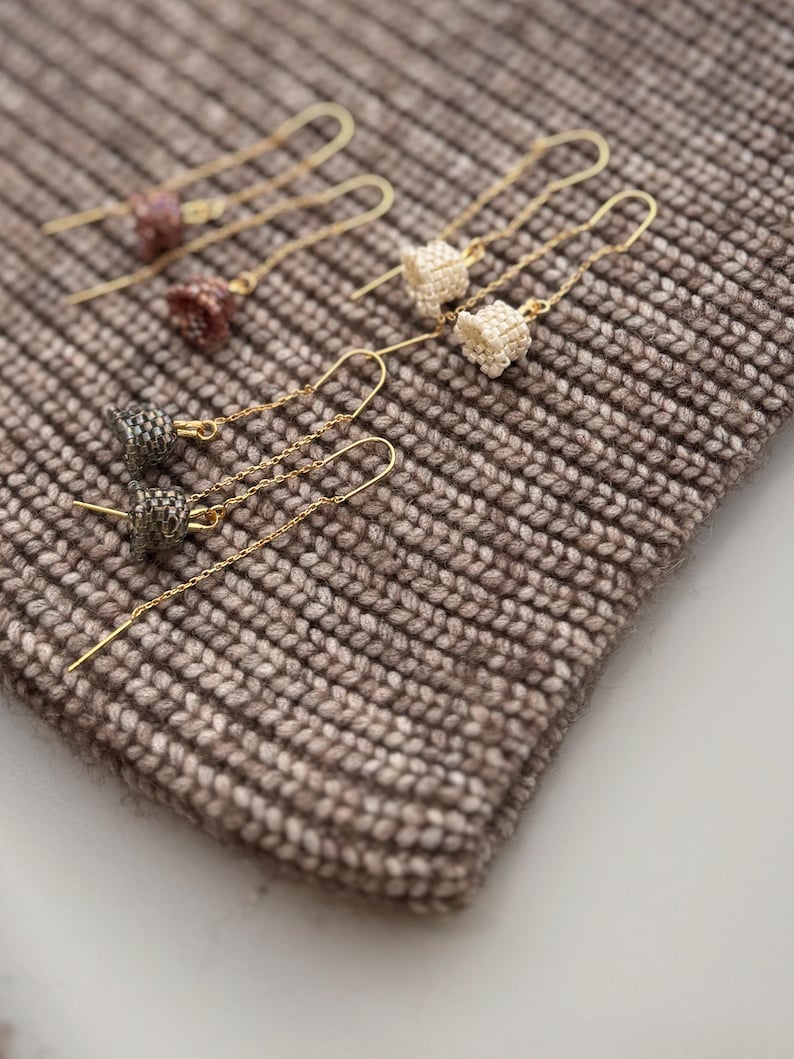 May include: A set of six gold dangle earrings with different colored beaded charms. The earrings are displayed on a brown knitted surface.