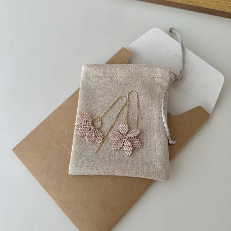 May include: A pair of delicate pink flower earrings with gold chain dangles, packaged in a small beige pouch.