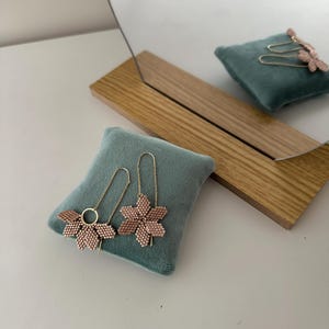 May include: A pair of delicate gold earrings with pink flower-shaped charms. The earrings are displayed on a teal velvet cushion.