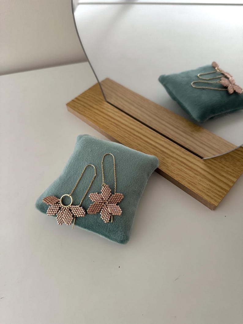 May include: A pair of delicate gold earrings with pink flower-shaped charms. The earrings are displayed on a teal velvet cushion.