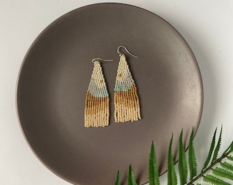 May include: A pair of long, beaded earrings with a gradient of beige, light blue, and gold. The earrings are hanging from a silver hook.