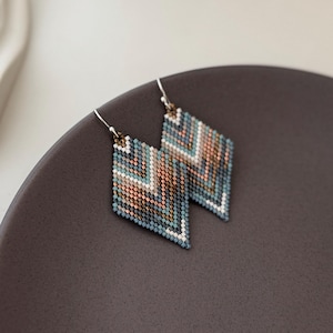 Könnte beinhalten: Ein Paar geometrisch gemusterte Ohrringe mit Chevron-Design. Die Ohrringe bestehen aus kleinen Perlen in Blautönen, Weiß und Kupfer.