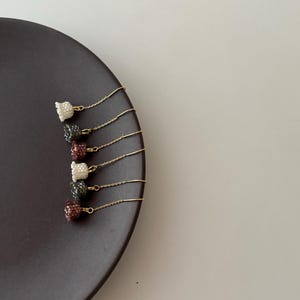 May include: Five pairs of gold chain earrings with small, beaded, bell-shaped charms in various colors, including white, gray, and burgundy. The earrings are displayed on a dark brown plate.