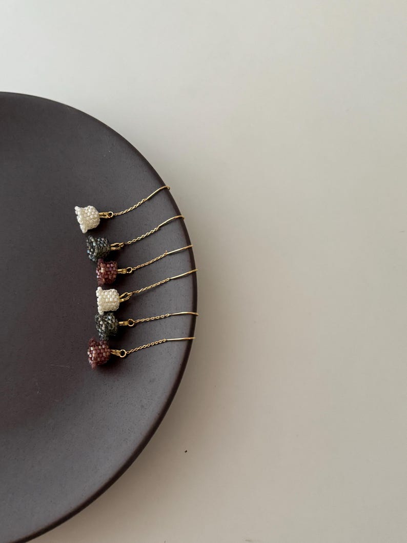 May include: Five pairs of gold chain earrings with small, beaded, bell-shaped charms in various colors, including white, gray, and burgundy. The earrings are displayed on a dark brown plate.