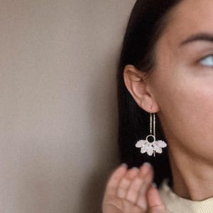 May include: A pair of long, delicate earrings with a gold chain and a small, circular charm. The earrings feature a fan-shaped design with multiple layers of small, white beads.