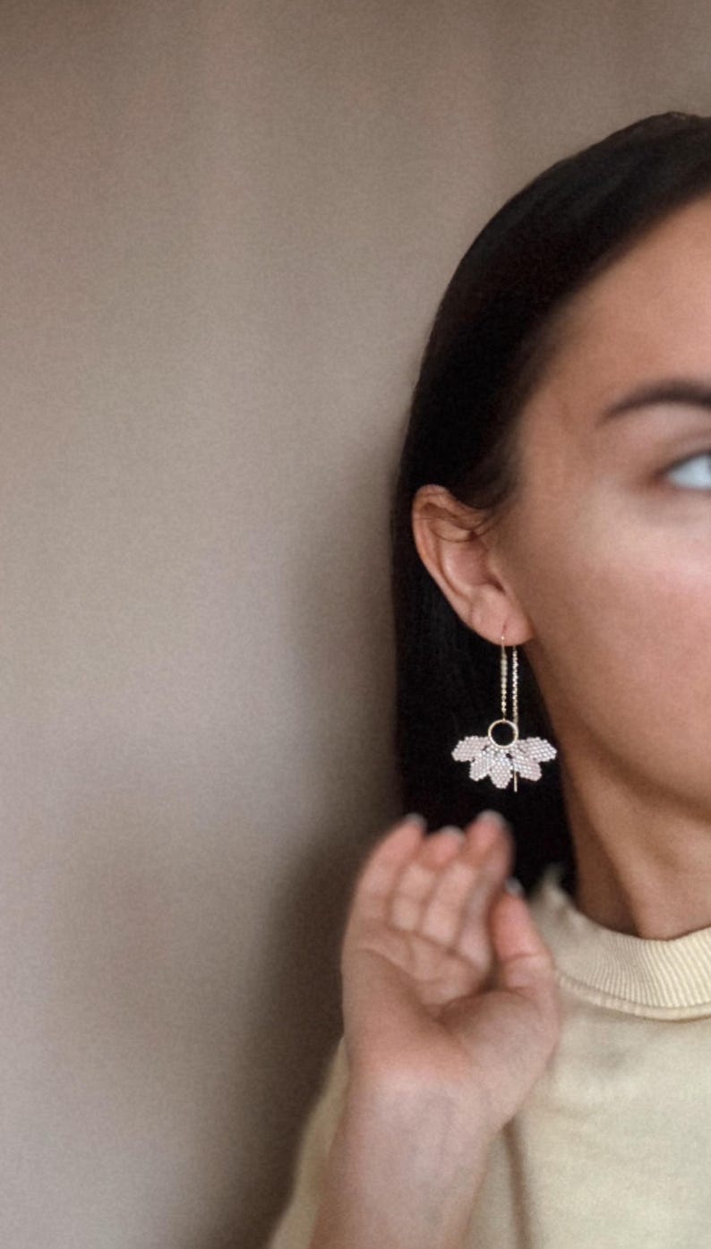 May include: A pair of long, delicate earrings with a gold chain and a small, circular charm. The earrings feature a fan-shaped design with multiple layers of small, white beads.