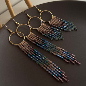 Puede incluir: Cuatro pares de pendientes de aro dorados con flecos de cuentas. Los flecos presentan un degradado de colores, incluyendo beige, azul y verde. Los pendientes se exhiben sobre una superficie gris oscuro.