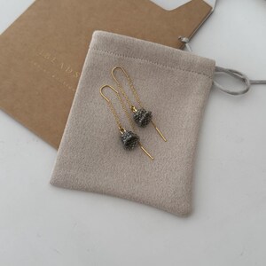 May include: A pair of gold dangle earrings with small gray beaded charms. The earrings are in a small beige pouch with the text "The Beads" on the pouch.