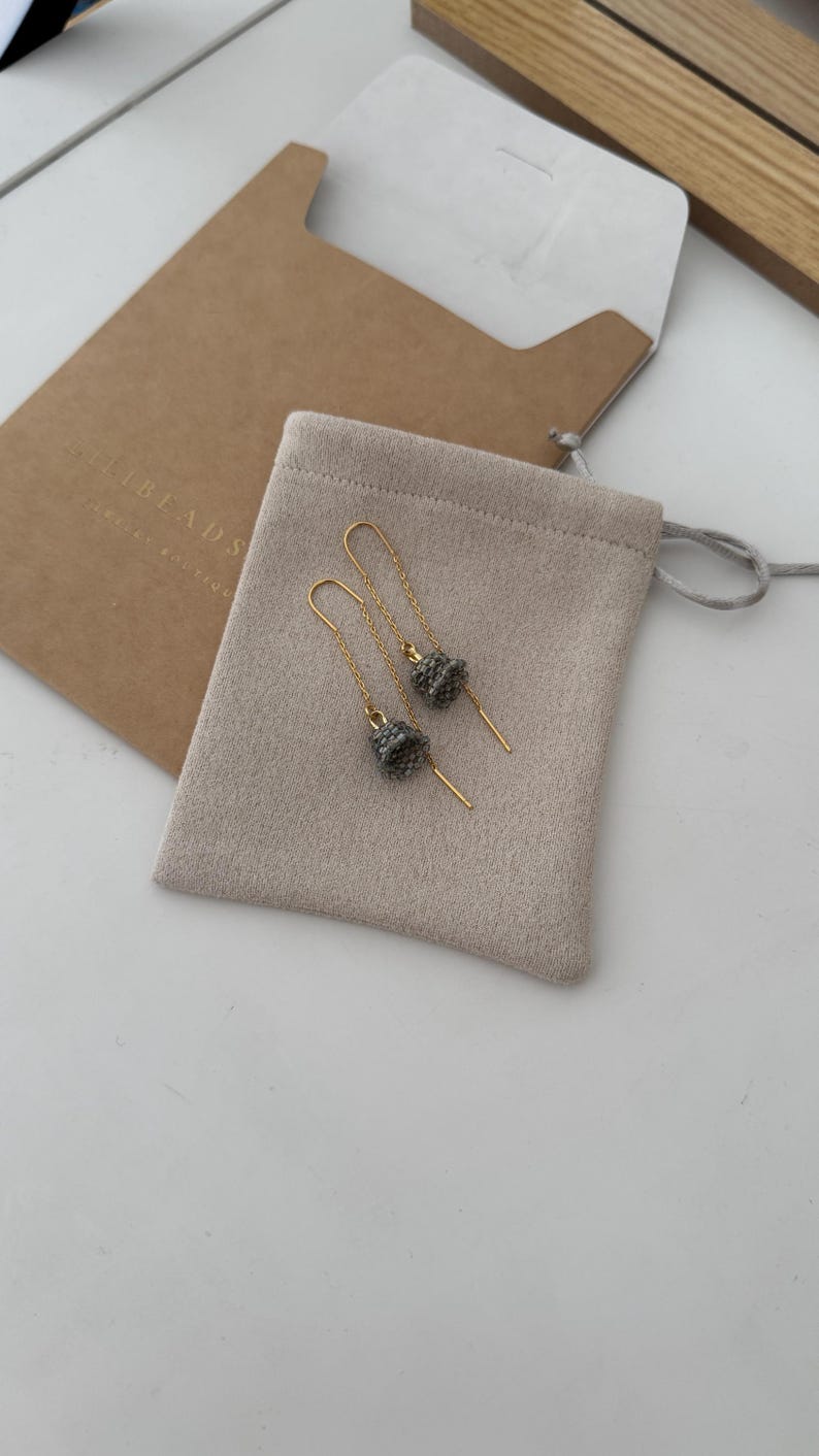 May include: A pair of gold dangle earrings with small gray beaded charms. The earrings are in a small beige pouch with the text "The Beads" on the pouch.