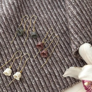 May include: Three pairs of dangle earrings with small, beaded, bell-shaped charms. The earrings are on a brown and white striped fabric background. The charms are in shades of gray, pink, and white.
