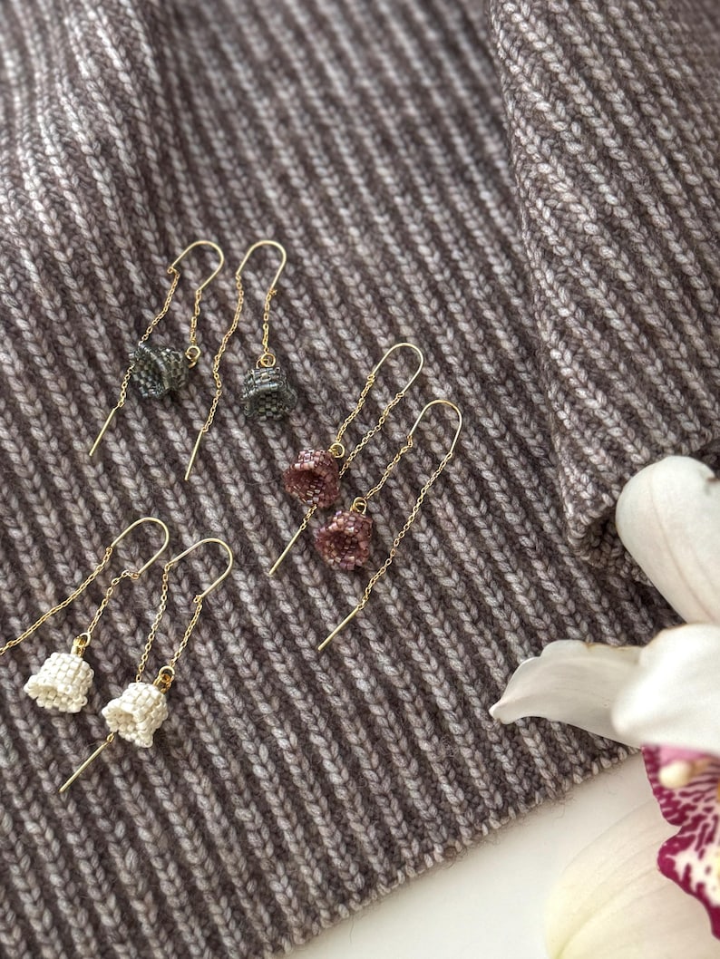 May include: Three pairs of dangle earrings with small, beaded, bell-shaped charms. The earrings are on a brown and white striped fabric background. The charms are in shades of gray, pink, and white.